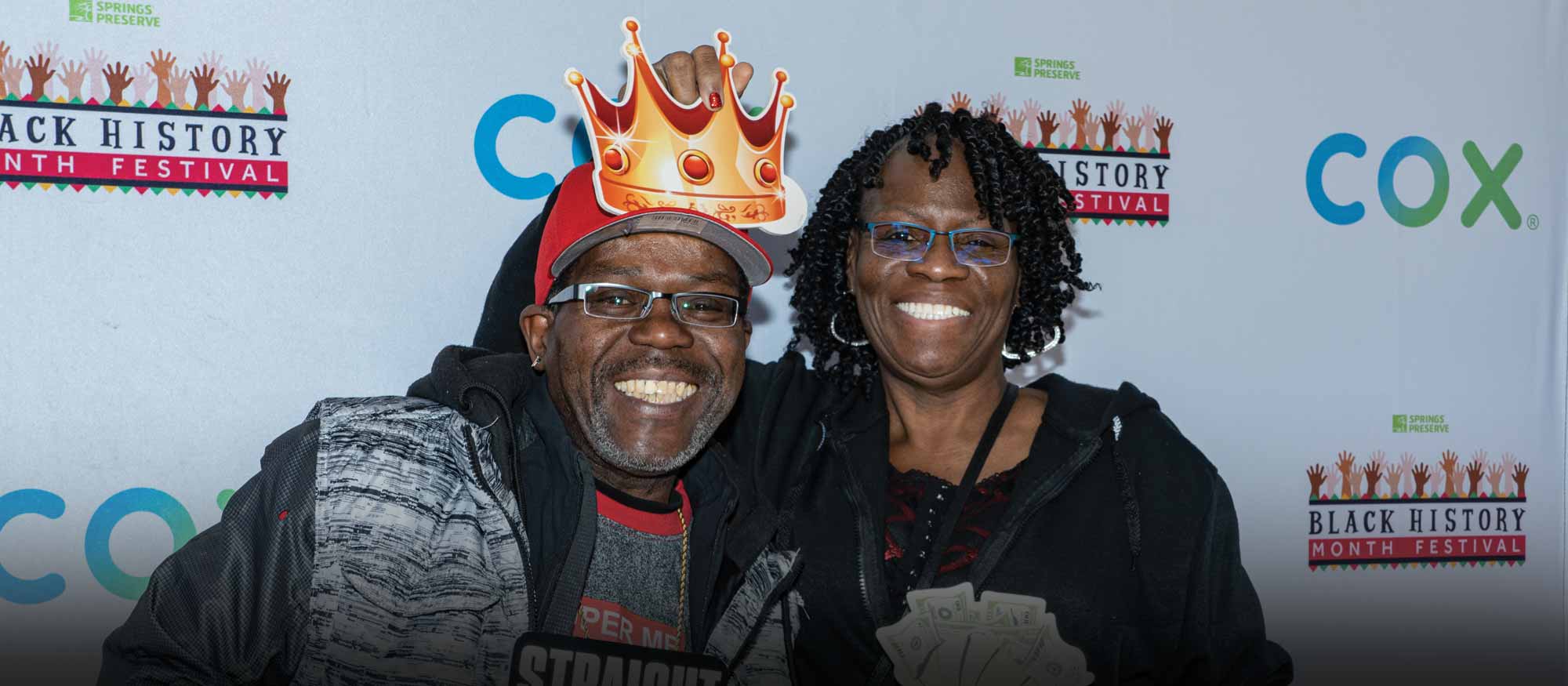 Two individuals smiling at a Black History Month Festival event sponsored by Cox, holding playful props.